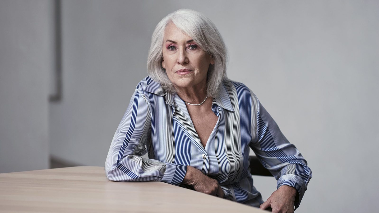 Judith Neilson wears a blue blouse. She is pictured sitting at a table with one hand resting on it and the other on her hip. She is looking at the camera and her hair is down.