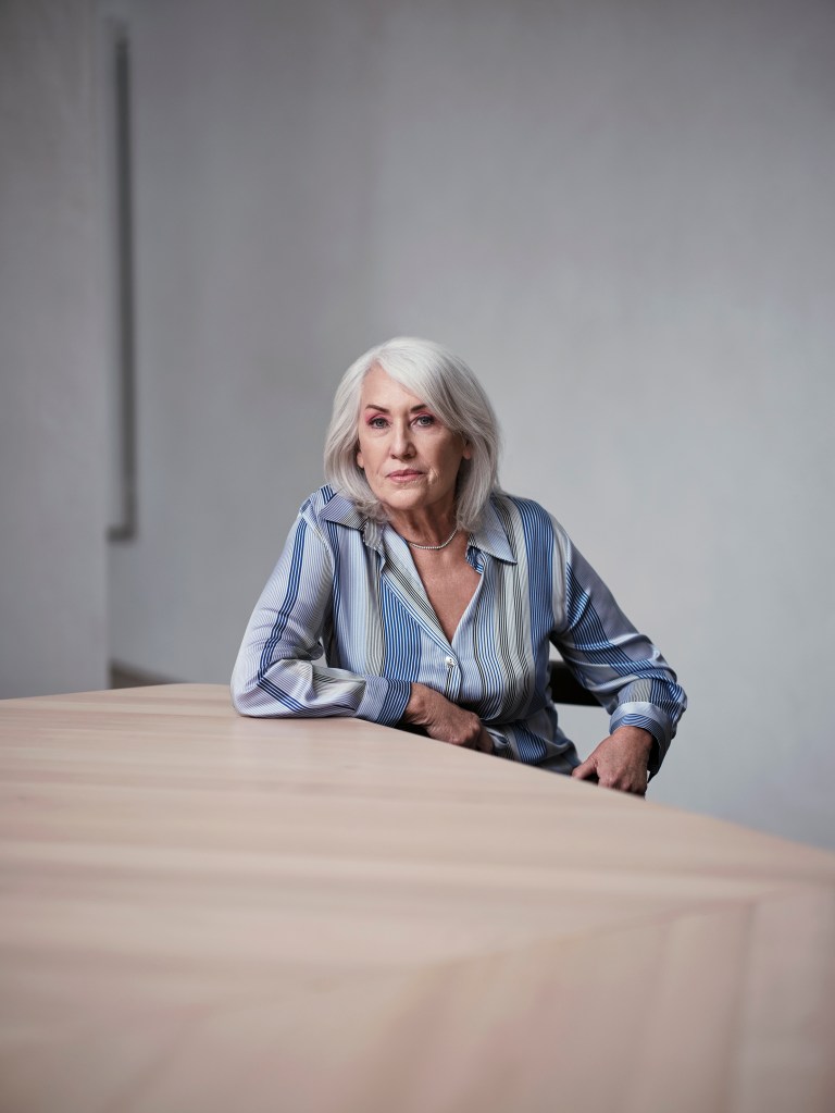 Judith Neilson wears a blue blouse. She is pictured sitting at a table with one hand resting on it and the other on her hip. She is looking at the camera and her hair is down.