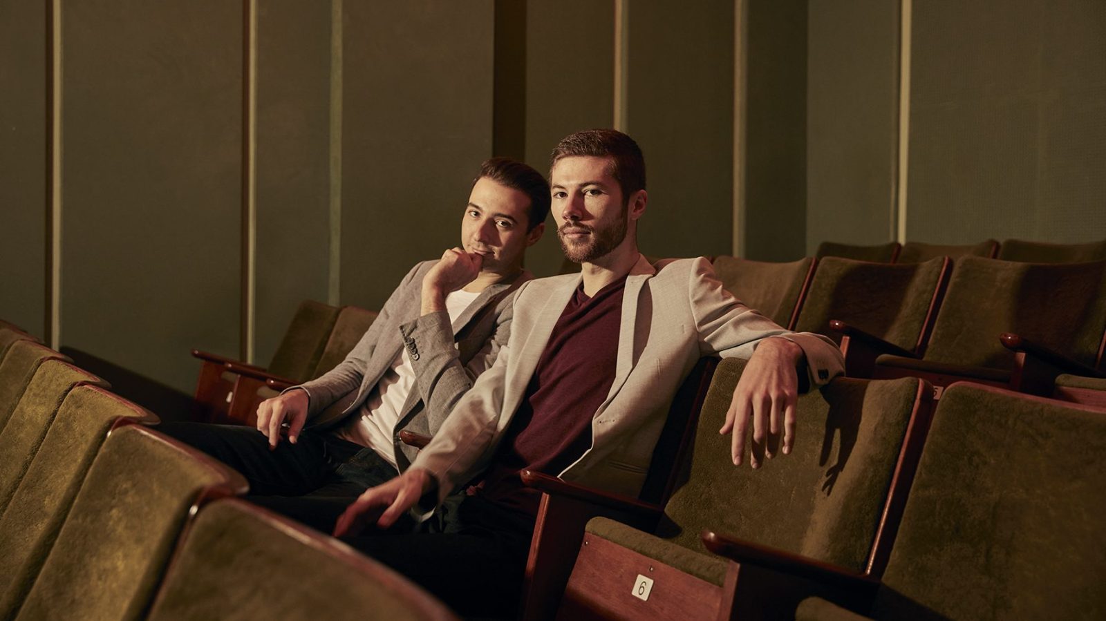 Alex Harper (left) and Angus Goldman (right) are sitting in a movie theatre. They are both wearing blazers with t-shirts underneath. They are looking at the camera.