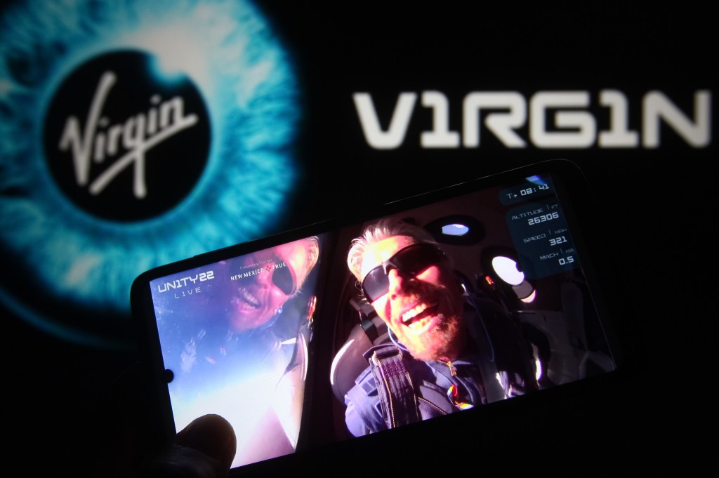 In this photo illustration, British billionaire Richard Branson is seen on a fragment of a Virgin Galactic Unity 22 Spaceflight Livestream Youtube video displayed on a smartphone with the Virgin Galactic logo in the background.  | Photo Illustration by Pavlo Gonchar/SOPA Images/LightRocket via Getty Images